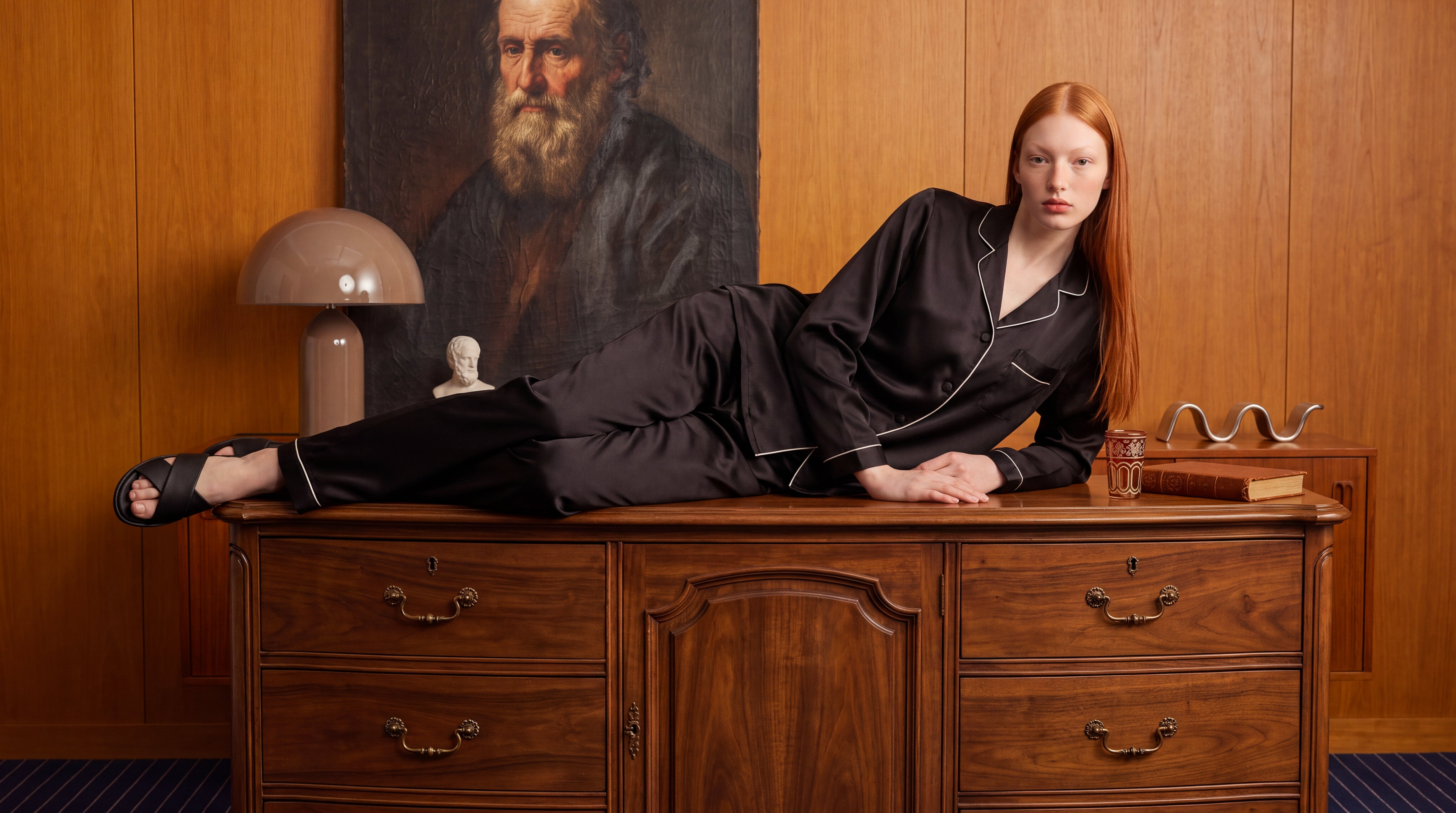 Woman in black pajamas sitting on a wooden dresser with a painting of a bearded man in the background.